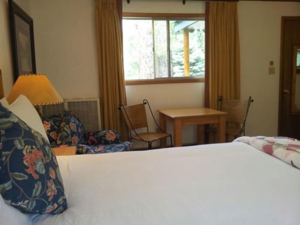 A hotel room with a bed, a table, and two chairs. There is a window with curtains, a framed picture on the wall, and a door.