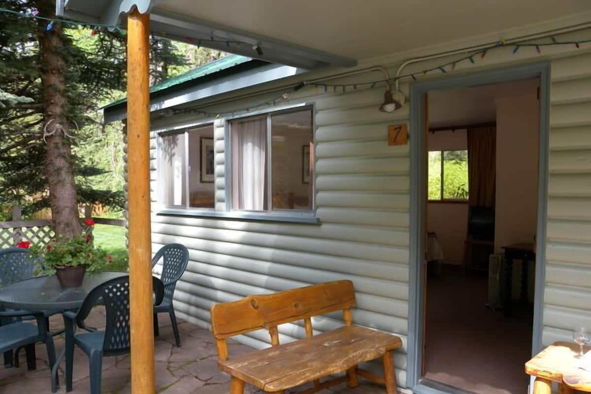 A wooden bench outside a cabin with a patio table and chairs, a potted plant, and a door leading to an interior room.