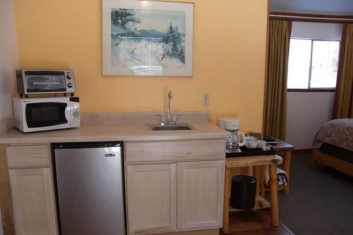 A compact kitchen with a microwave, toaster, and refrigerator. There's a sink, a small table with a coffee maker, and a framed picture on the wall.