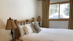 Una cama bien hecha con sábanas blancas y dos almohadas, flanqueada por dos mesitas de noche con lámparas, en una habitación con ventana y cortinas.
