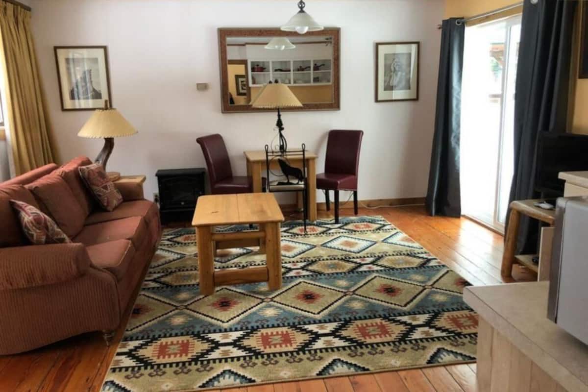 A cozy living room with a sofa, a wooden table, and chairs. There is a patterned rug on the floor, and a mirror on the wall.