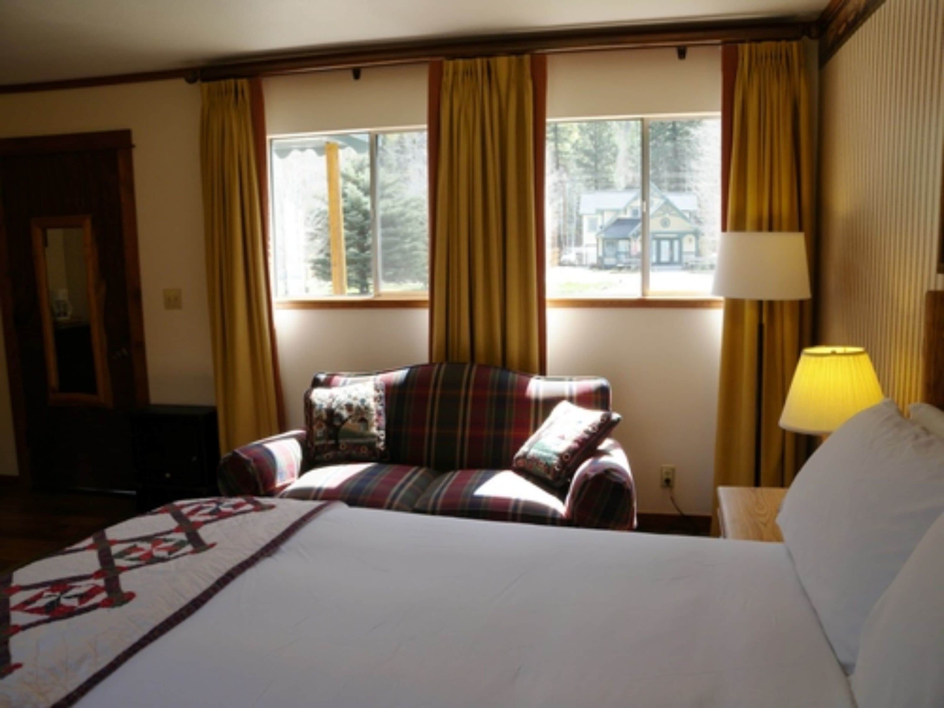 A hotel room with a bed, a plaid armchair, a window with curtains, and a lamp.
