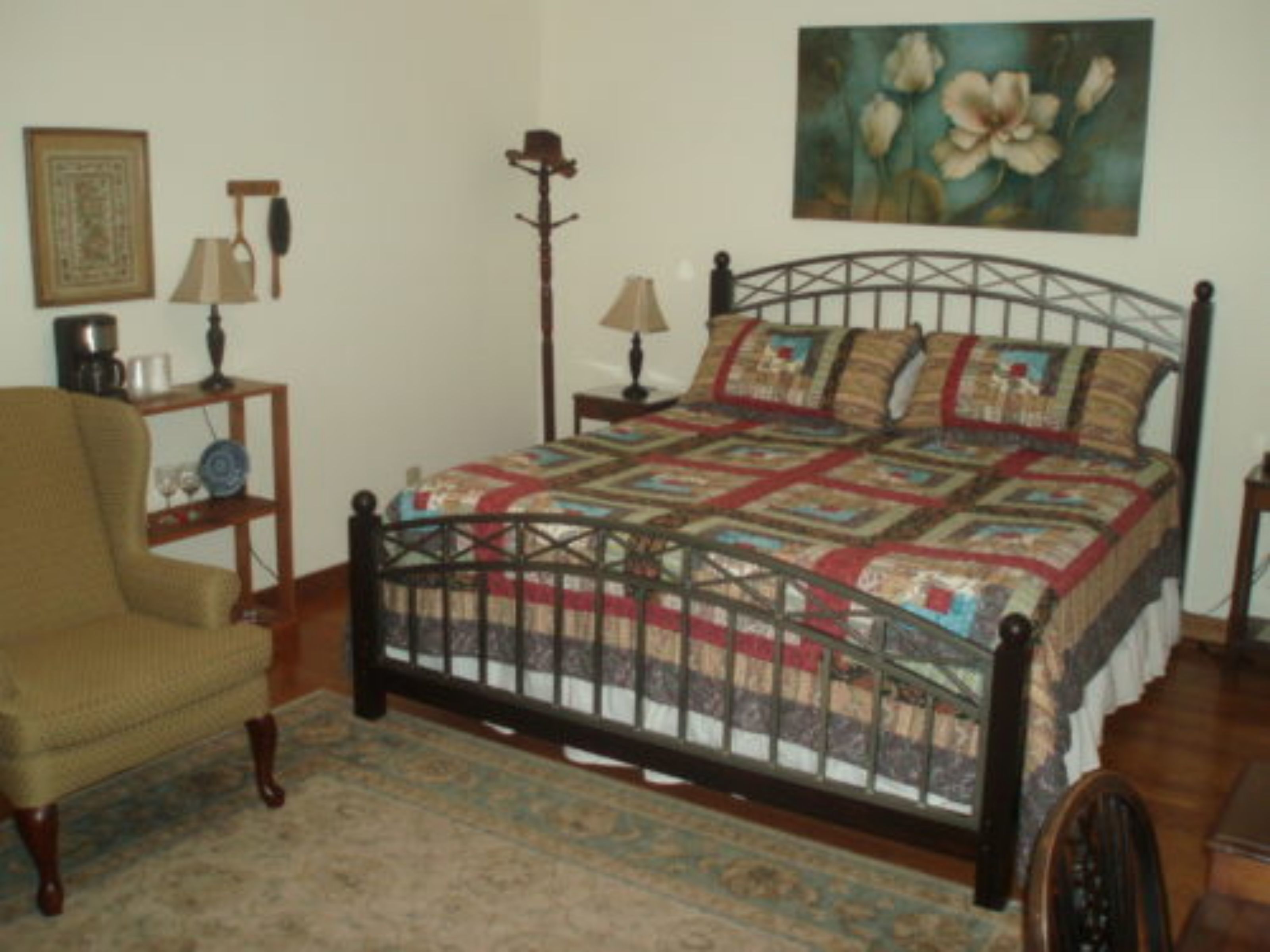 Un dormitorio con una cama, una silla, una lámpara, un cuadro y una mesita.