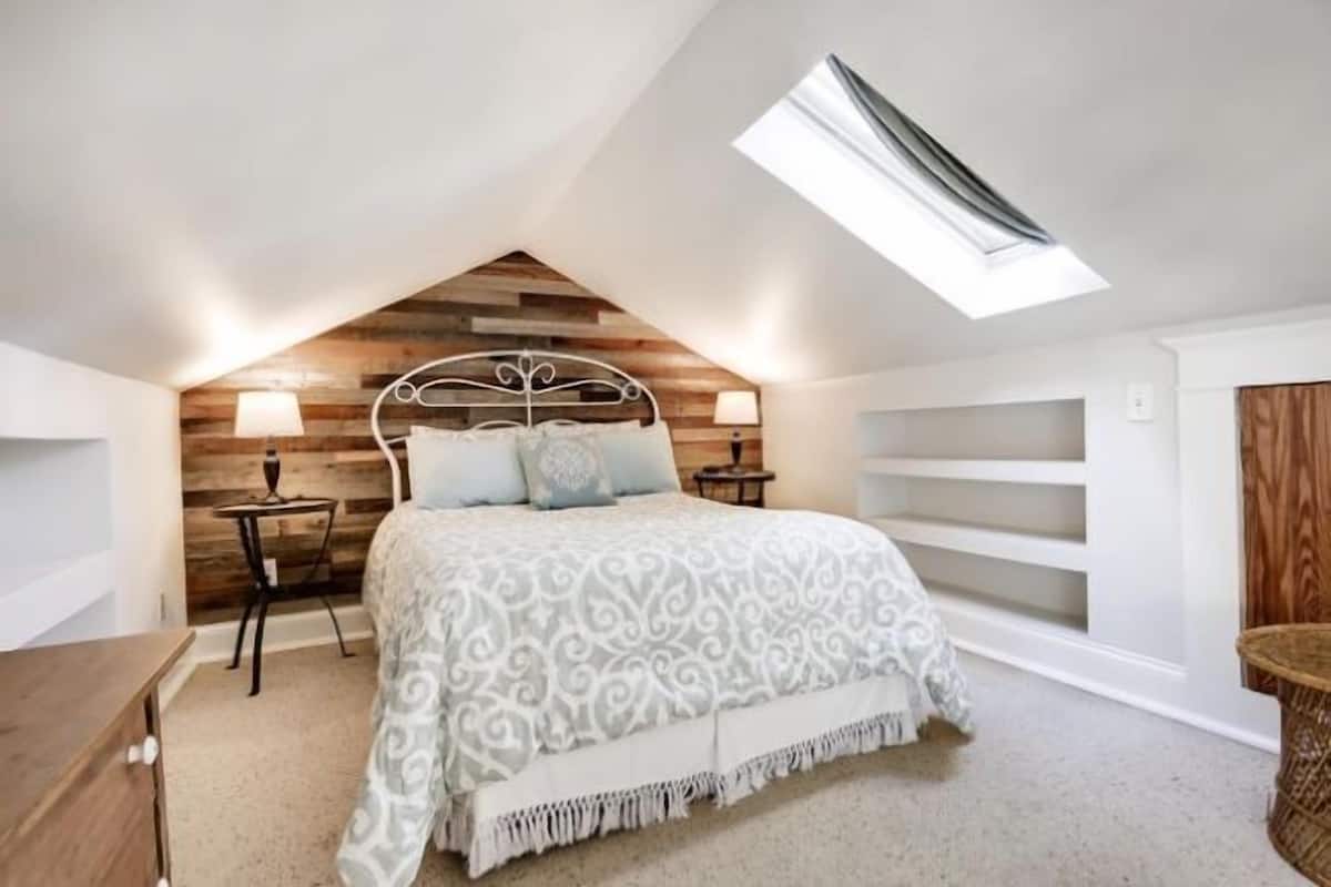 A bedroom with a sloped ceiling, a skylight, a bed with patterned bedding, two bedside tables with lamps, and a wooden headboard.
