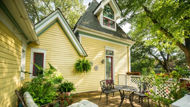 Une maison jaune avec un porche, une terrasse couverte et un petit jardin agrémenté de plantes et d’un banc.