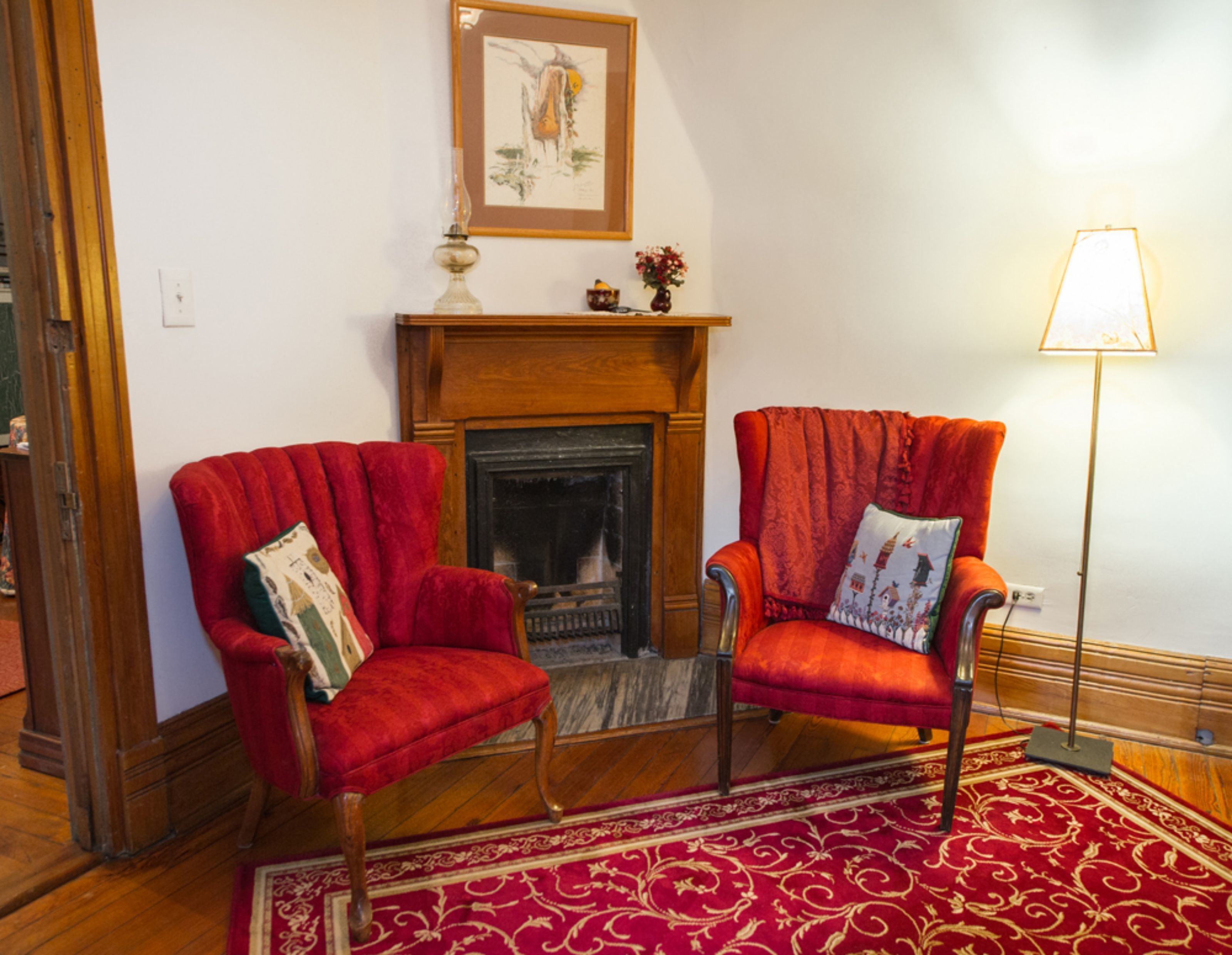 A room with two red armchairs, a fireplace, a framed picture above the mantel, and a floor lamp.
