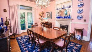 Une salle à manger avec une grande table en bois, des chaises et un tapis à motifs floraux. On y trouve un lustre, une cheminée et une fenêtre avec des rideaux.