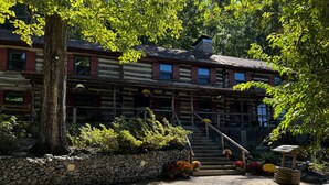 A log cabin with a stone foundation, a wooden staircase leading to a porch, and a gravel driveway.