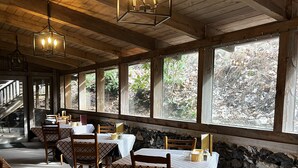 A restaurant with wooden beams, stone walls, and large windows overlooking a forest. Tables are set with checkered tablecloths and chairs.
