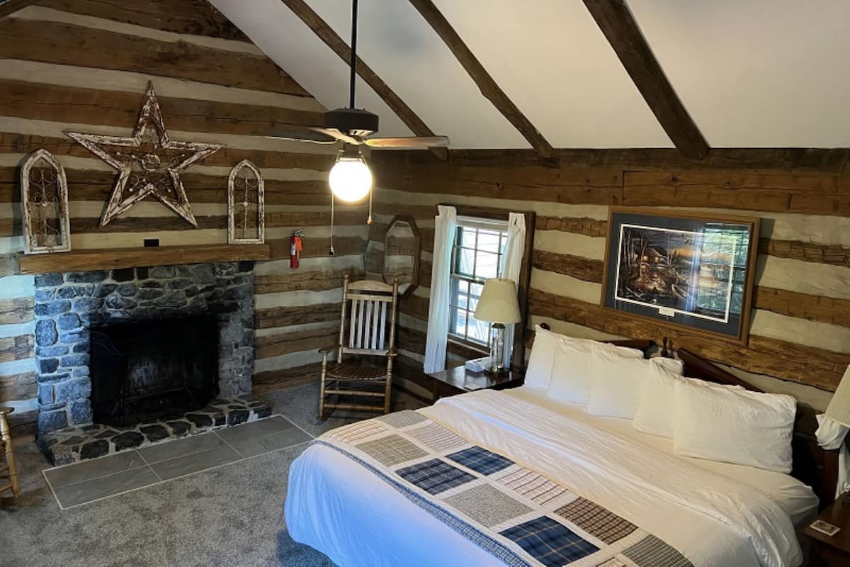 A log cabin bedroom with a stone fireplace, a bed with a quilt, a wooden chair, and a ceiling fan.