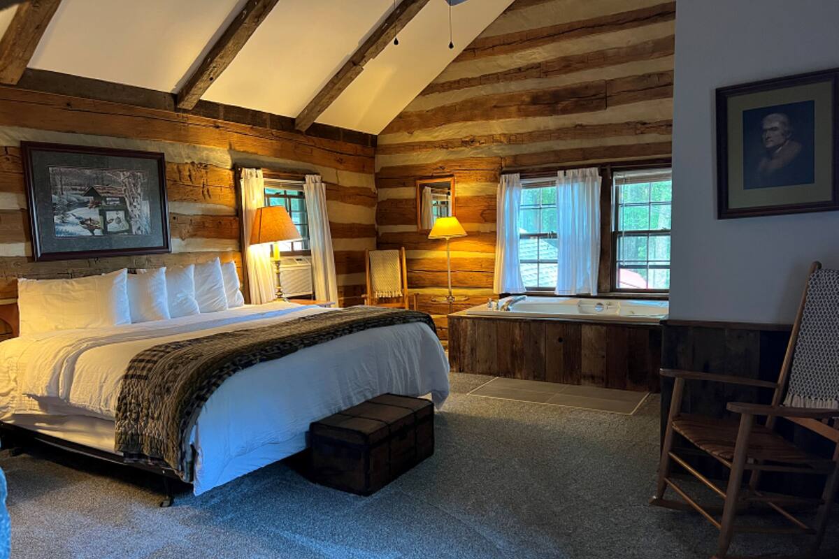 A log cabin-style bedroom with a large bed, a wooden bench, a chair, a desk, and a window with curtains.