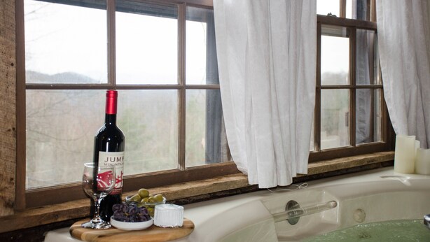 A room with a large window, a bathtub, a wine bottle, and a glass of wine on a wooden tray.