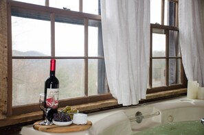 A room with a large window, a bathtub, a wine bottle, and a glass of wine on a wooden tray.