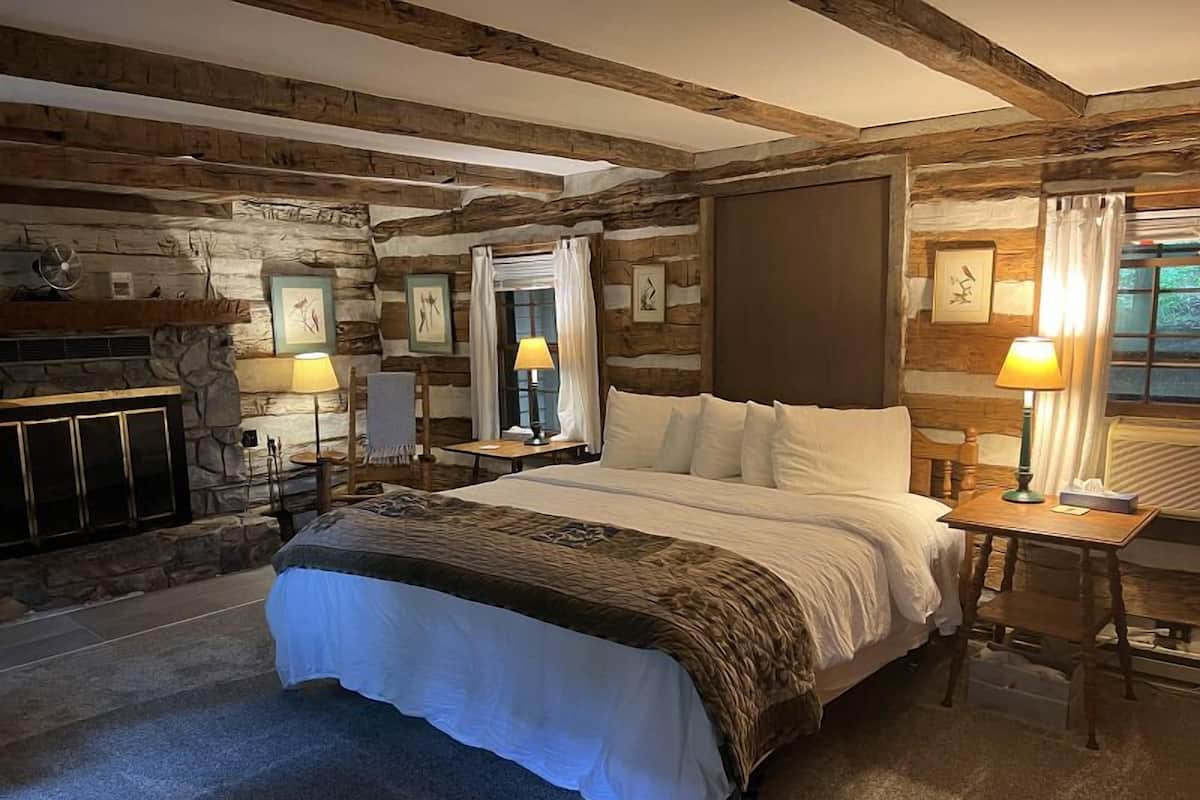 A cozy log cabin-style bedroom with a large bed, two bedside tables with lamps, a fireplace, and stone walls.