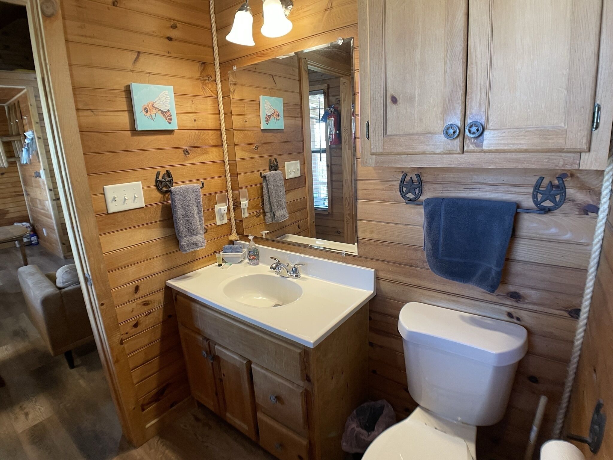 Une salle de bain rustique, avec une cabine en bois, un lavabo blanc, un miroir et des toilettes. On y trouve également des armoires en bois et une corde accrochée au mur.