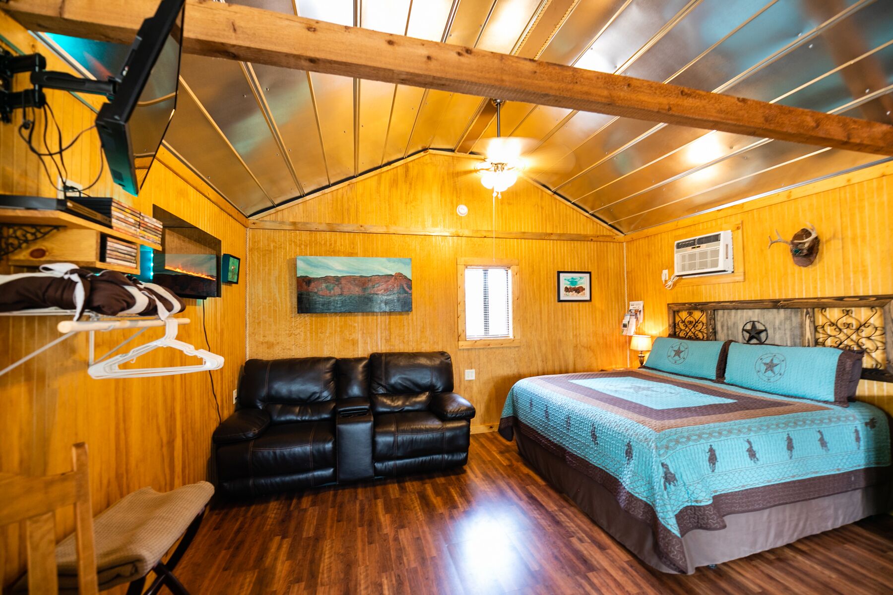 Una habitación acogedora con techo de madera, una cama con colcha azul y marrón, un sofá de cuero negro y suelo de madera.