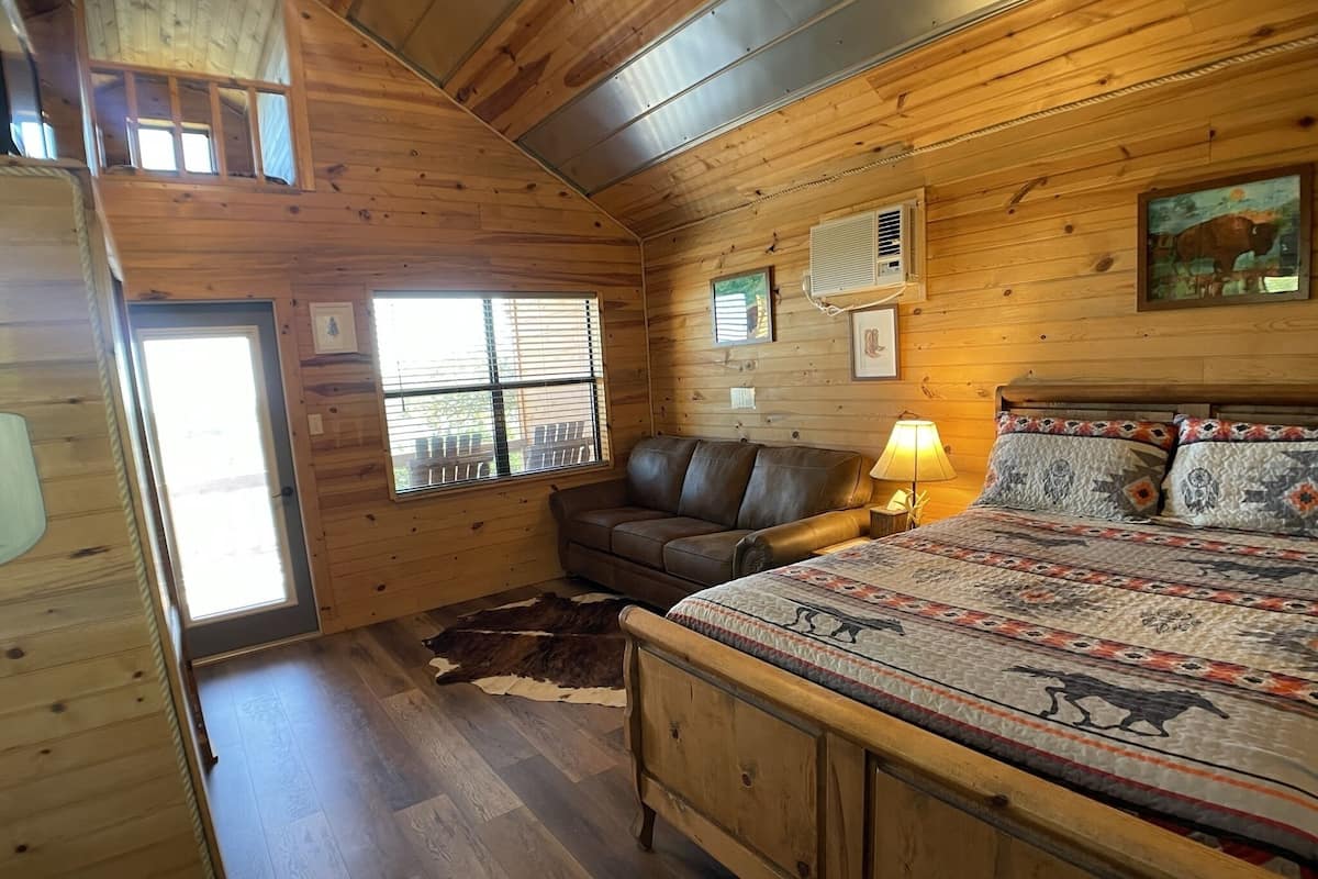 A cozy cabin room with a wooden interior, a leather sofa, a bed with patterned bedding, and a cowhide rug.