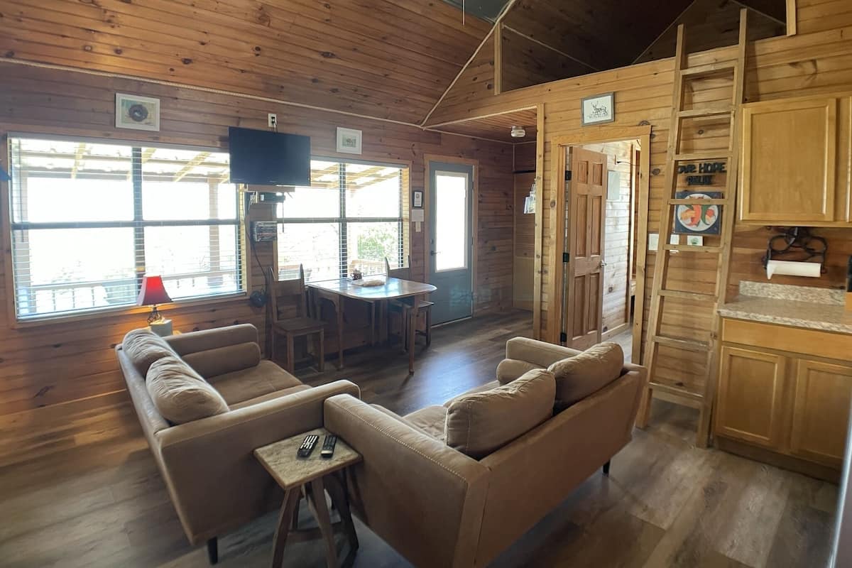 A wooden cabin interior with a sofa, a small table, a TV, and a kitchen area.