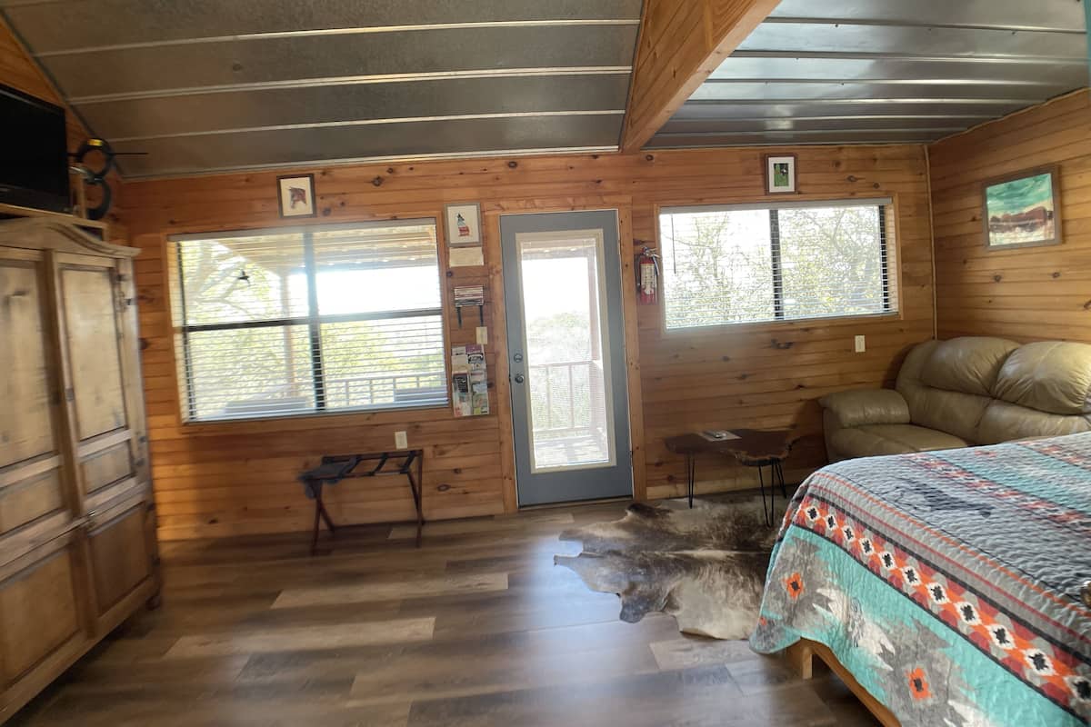 A room with wooden walls, a bed with a patterned blanket, a leather couch, a wooden cabinet, and two windows with blinds.