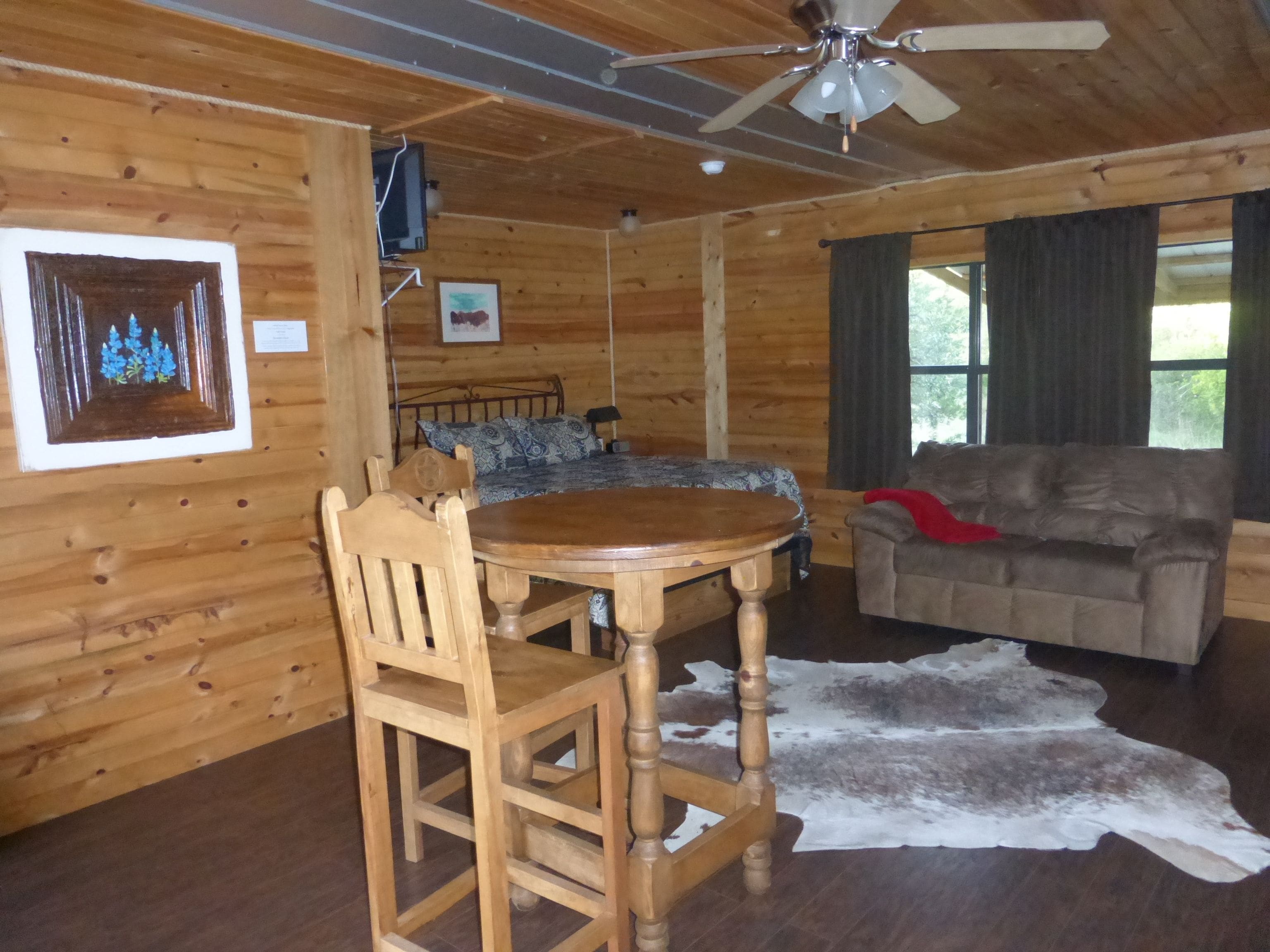 A wooden cabin interior with a dining table, chairs, a sofa, a bed with patterned bedding, and a ceiling fan.