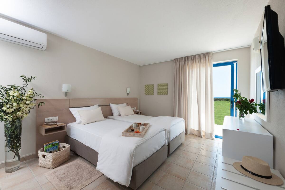 A hotel room with two beds, a TV, a desk, a window with curtains, a plant, and a tray with breakfast items.