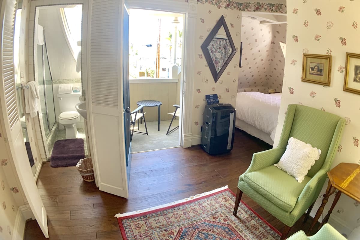 Quarto com cama, poltrona verde, mesa pequena, escrivaninha e banheiro visível por uma porta aberta.