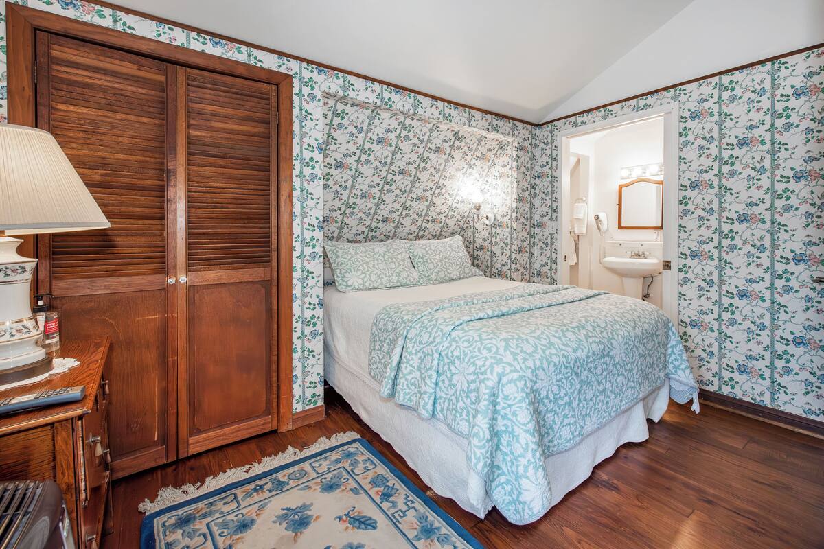 Quarto com guarda-roupa de madeira, cama com colcha estampada, mesa de cabeceira com abajur e banheiro com espelho e pia.
