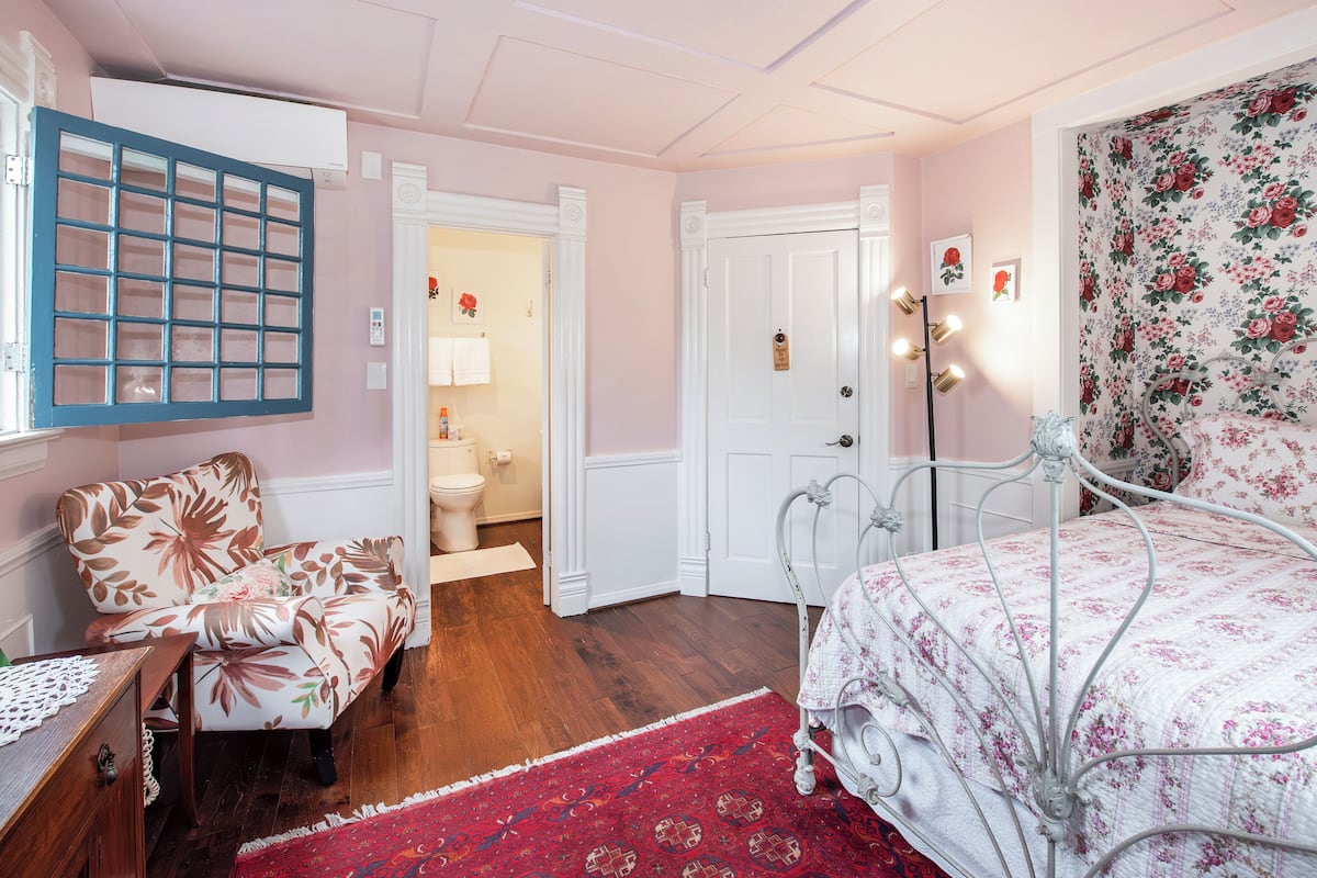 Quarto com uma cadeira estampada em flor, estrutura de cama em metal, tapete vermelho e porta branca que dá para um banheiro.