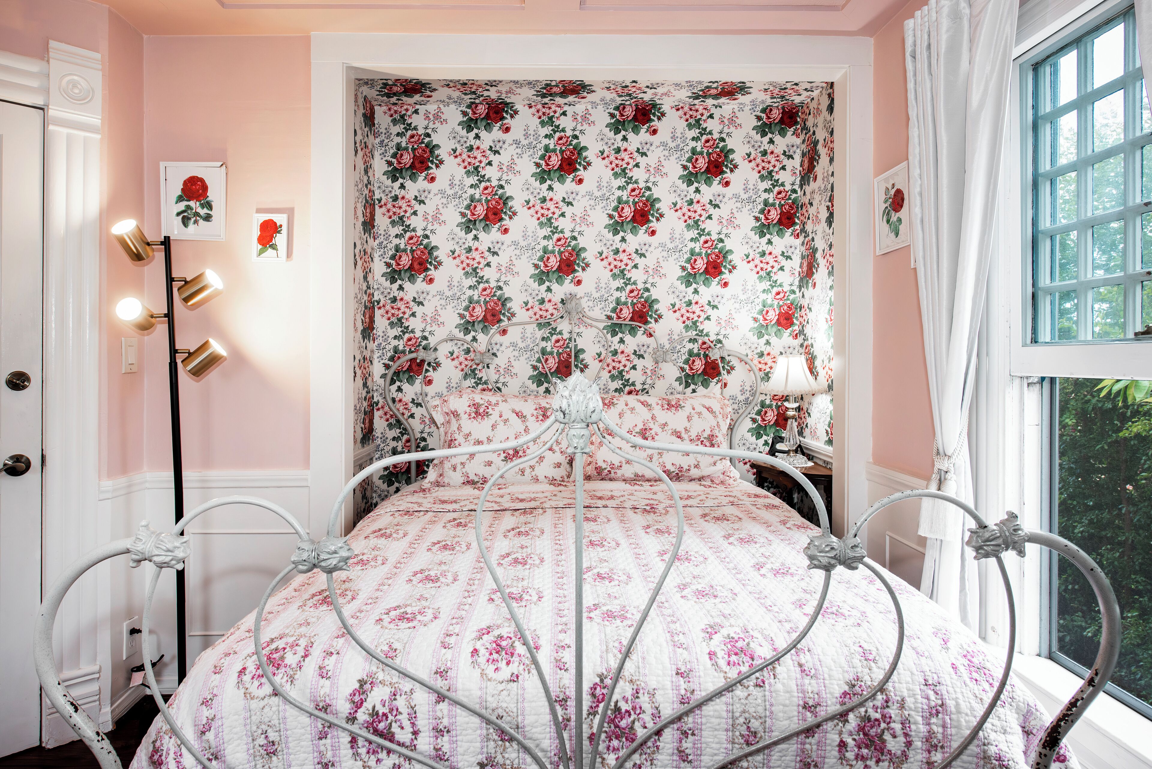 Una camera da letto con una coperta a motivi floreali, una struttura letto in metallo bianco, un comodino con una lampada e una finestra con tende.