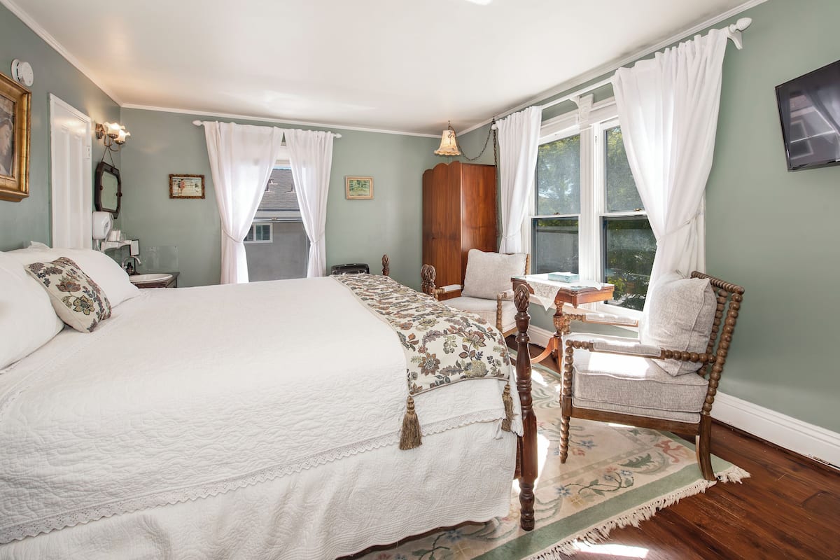 Quarto com uma cama grande, uma cadeira de madeira, uma escrivaninha e uma janela com cortinas.
