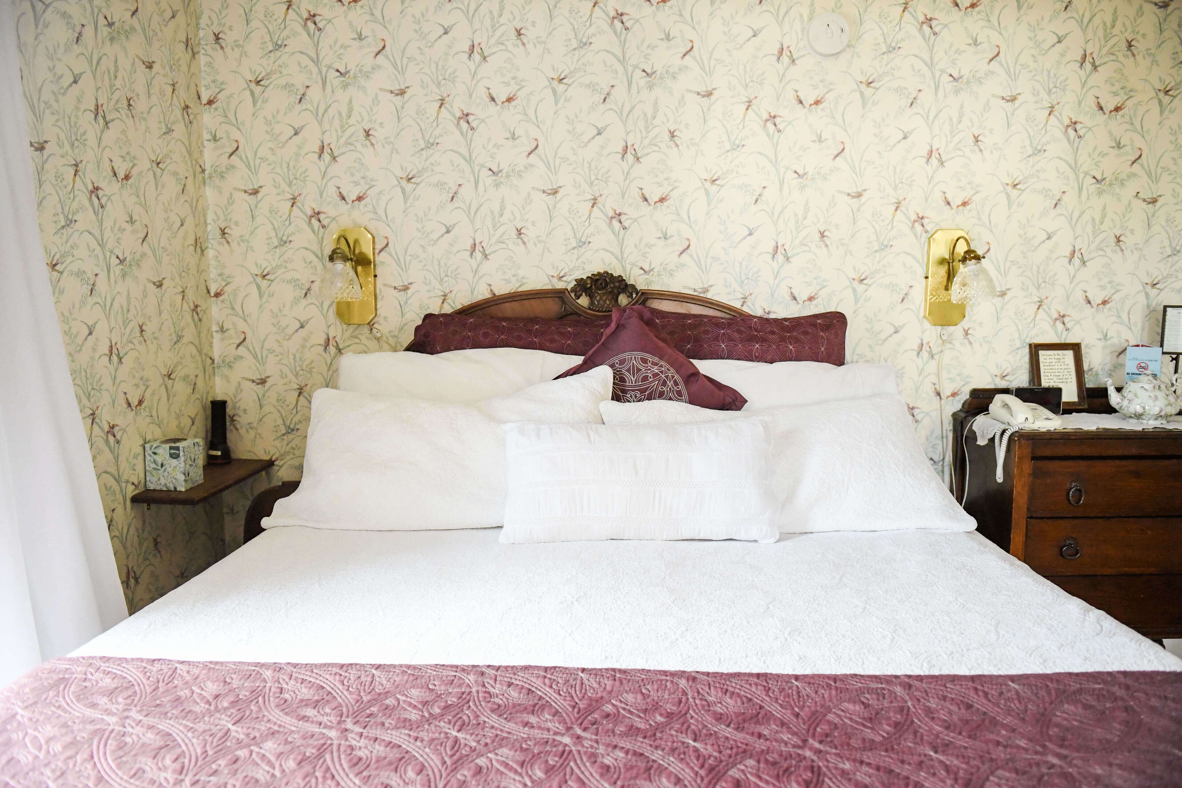 Una cama bien hecha con ropa de cama blanca y un cabecero decorativo, una mesita de noche con lámpara, y una lámpara de pared.