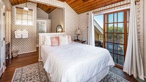 A bedroom with a large bed, a wooden ceiling, a window with curtains, and a view of greenery outside. - Bath Street Inn (Santa Barbara)
