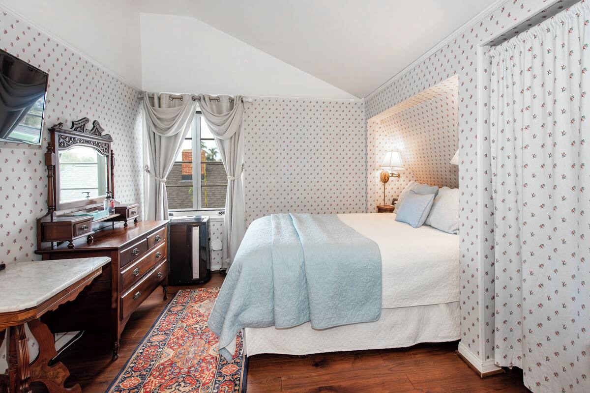 Quarto com cama, cômoda, escrivaninha, espelho, janela com cortinas e tapete estampado.