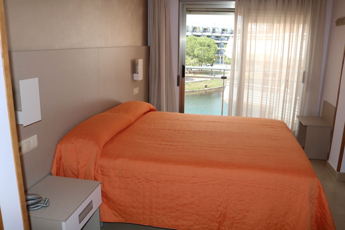 A hotel room with a bed, a bedside table with a telephone, and a view of a building and a body of water through the window.