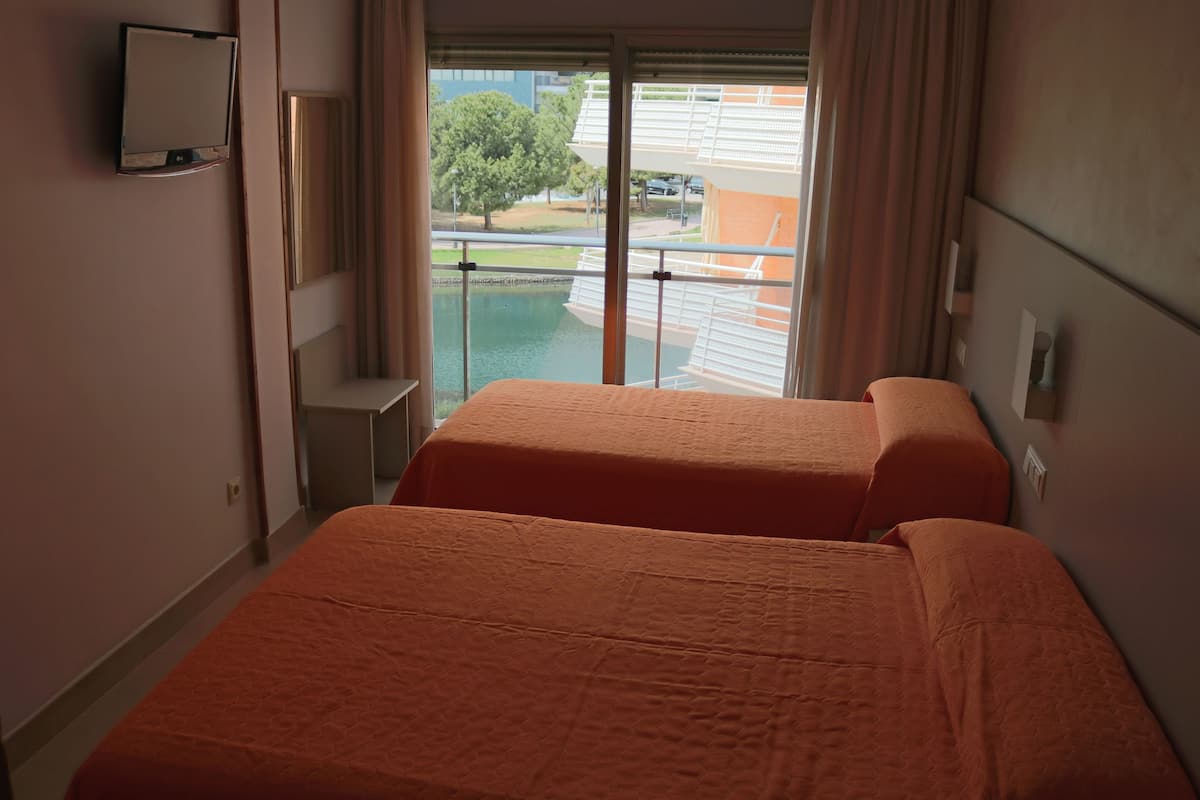 A hotel room with two beds, a TV mounted on the wall, and a view of a swimming pool and buildings outside.
