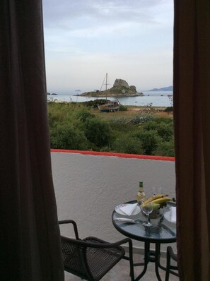 A balcony with a table set for two, overlooking a coastal area with boats and a rocky island.