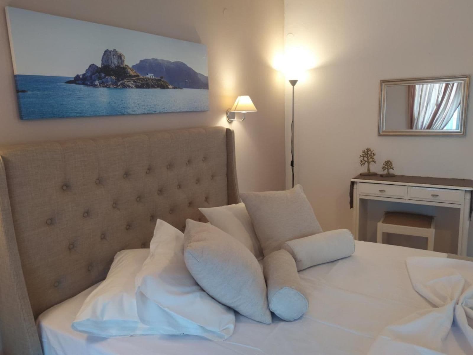 Cama bem arrumada com cabeceira, luminária de cabeceira, mesinha com espelho e pintura de uma paisagem costeira na parede.