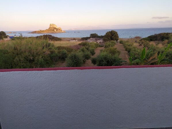 A coastal landscape with a prominent rock formation, a path leading to a beach, and sparse vegetation. - Kaluva Studios (Kos)