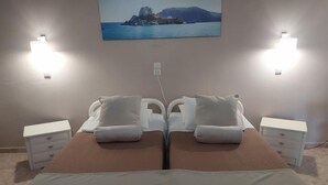 Una cama doble con dos mesitas de noche, un cuadro de un paisaje costero e iluminación en la pared.