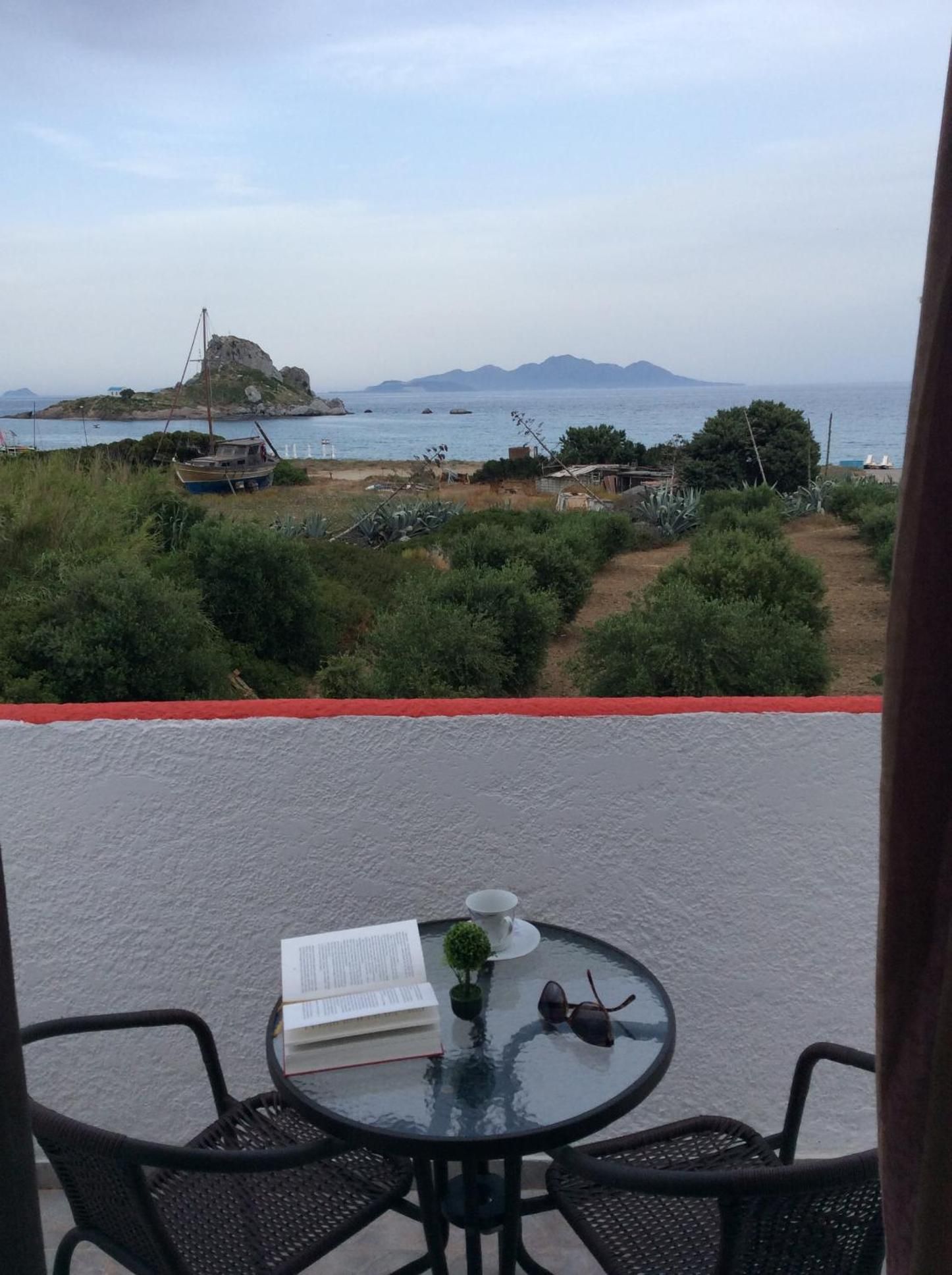 Een balkon met een tafel, stoelen en een boek, met uitzicht op een kustlandschap met begroeiing en een verre zee.