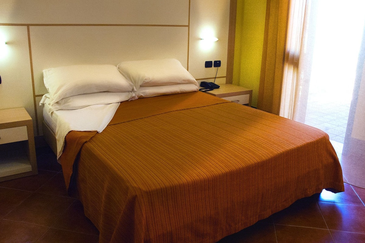 Una cama bien hecha con un edredón marrón, dos mesitas de noche con lámparas, y vistas a un suelo de baldosas y una cortina.
