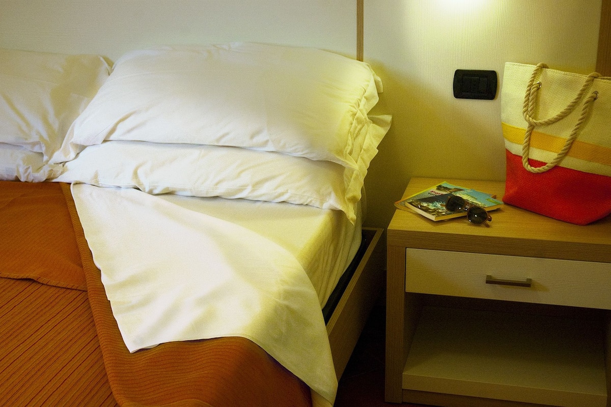 Una cama bien tendida con sábanas blancas, una mesita de noche de madera con un bolso a rayas rojo y amarillo, y un libro y gafas de sol sobre la mesita.
