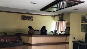 A reception area with a wooden reception desk, two seated individuals, a framed picture on the wall, and a window with curtains. - Indraprastham Tourist Home (Kottayam)