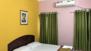 A hotel room with a bed, a picture on the wall, green curtains, and an air conditioning unit. - Indraprastham Tourist Home (Kottayam)