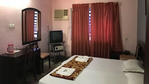 A hotel room with a bed, a desk with a chair, a television, and a window with curtains. - Indraprastham Tourist Home (Kottayam)