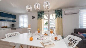 Une cuisine moderne avec des meubles blancs, une machine à café et un panier de fruits. On y trouve également un coin repas avec une table blanche, des chaises et un livre.