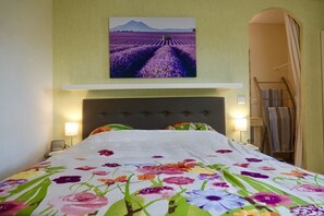 Et hotelværelse med en seng med blomstret sengetæppe, knægt, sengeborde med lamper og et billede af lavendelmarker over sengen.