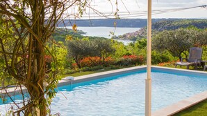 En swimmingpool med en pergola ovanför, omgiven av frodig grönska och med utsikt över en sjö och kullar.