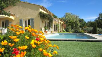 Une maison avec une piscine, entourée d’un jardin où poussent des fleurs jaunes et rouges.