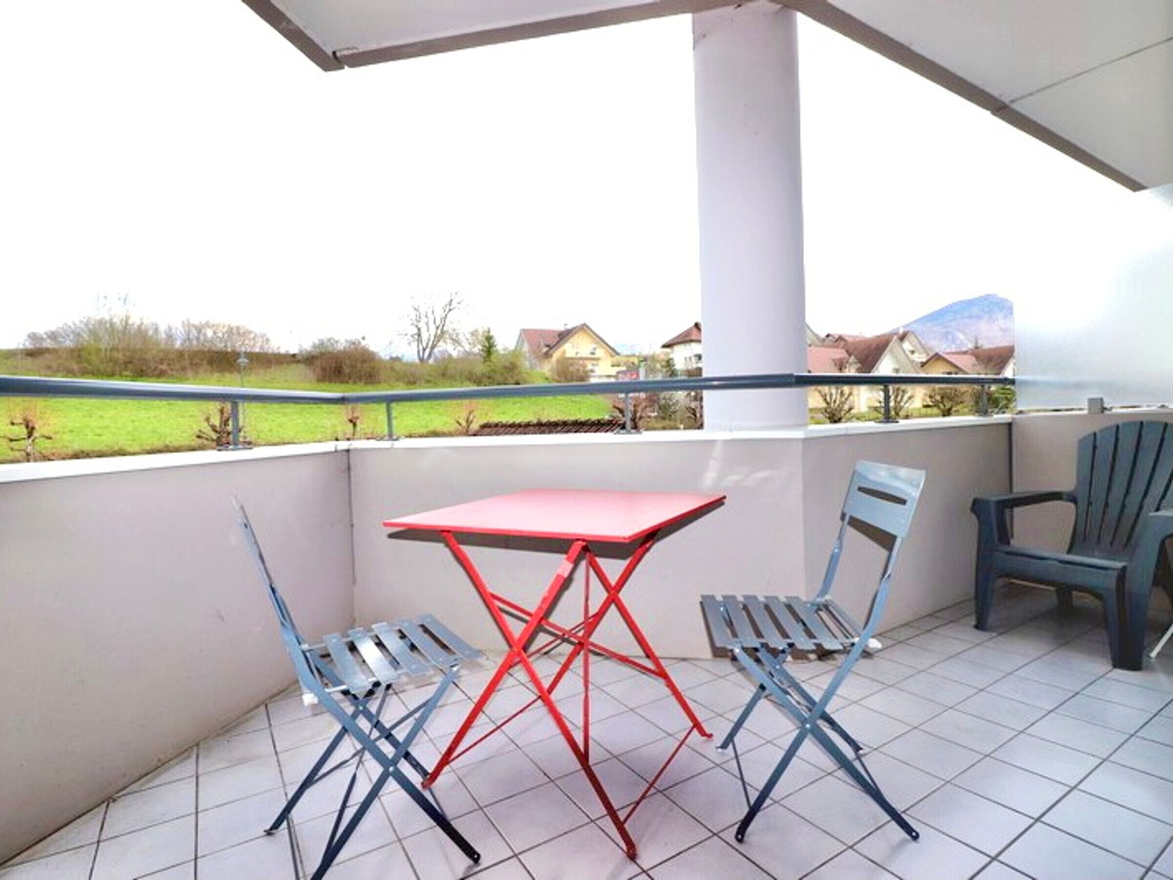 A balcony with a red table and two blue chairs, a white pillar, and a view of houses and greenery.